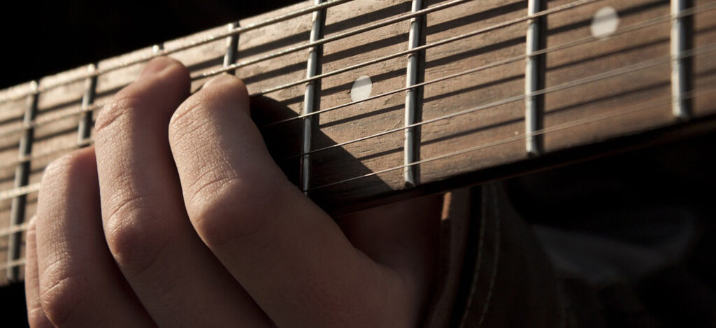 Close-up of a person's hand playing chords on a guitar neck, with strings and frets visible in natural lighting.